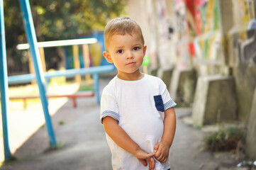 A cute blond boy in a white T-shirt stands modestly and looks into the lens with big eyes.