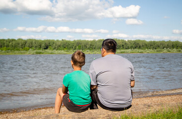 father and son sit on a log and look at the river, view from the back