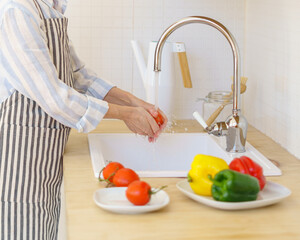 Unrecognizable senior woman grandmother in apron washing fresh vegetables for salad while enjoying cooking in modern light kitchen at home. Elderly woman preparing healthy food for family, vertical