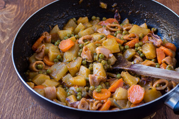 Squid with potatoes, peas and carrots cooked in a pan on wooden background