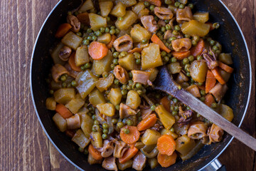 Squid with potatoes, peas and carrots cooked in a pan on wooden background
