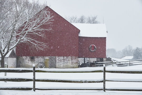 A architectural gem of a red barn with a green wreath photographed during a christmas time snowstorm