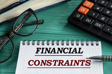 FINANCIAL CONSTRAINTS - words in a notepad on a wooden green background with a calculator, pen and glasses