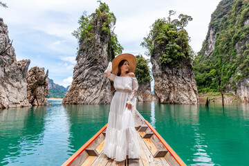 Fototapeta na wymiar Happy Young Female Tourist in Dress and Hat at Longtail Boat near Famous Three rocks with Limestone Cliffs at Cheow Lan Lake. Travel Woman Standing on Boat in Khao Sok National Park in Thailand