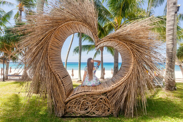 Back View of Tourist Woman Sitting in Photo Spot like Straw Heart with Tropical Palm Trees and Blue Sea with White Sandy Beach on Background. Nature and travel concept