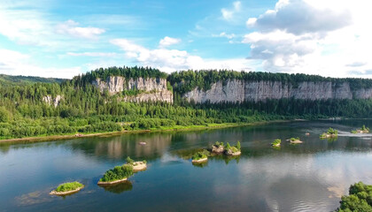 A huge rock mass stands like a wall along the river . Beautiful blue sky and shaggy clouds . Green forest along the river, small islands . Catamarans are rafted on the river .