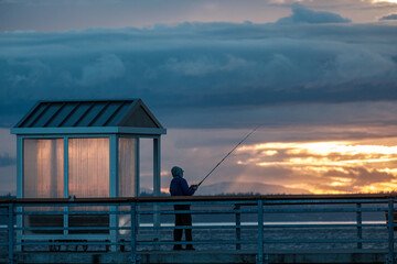 A silhouette of a person fishing off of a pier at sunset on an overcast day