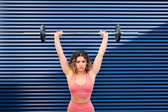 Female athlete lifting barbell in front of blue corrugated wall