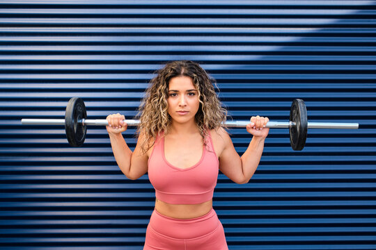 Young female athlete standing with barbell in front of blue wall