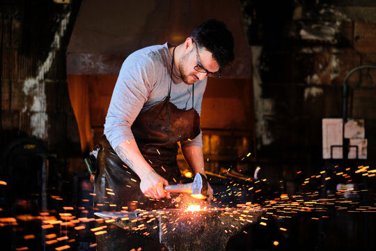 Young male blacksmith wearing eyeglasses forging with hammer at workshop