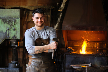 Smiling young male blacksmith in apron standing with hammer at workshop