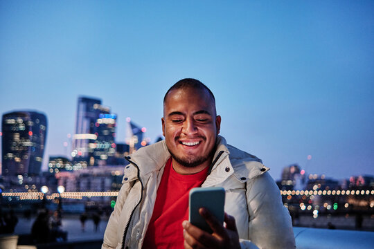 Smiling young man in padded jacket using smart phone in city at night