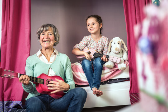 Smiling grandmother and granddaughter with guitar sitting in bedroom