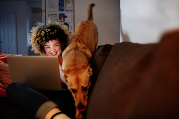 Cheerful young woman using laptop sitting with playful dog at home