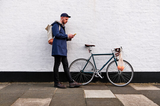 Man with bicycle using smart phone while leaning on wall