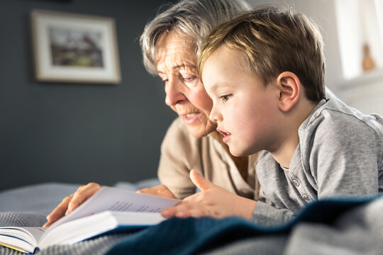 Grandmother reading book while lying by grandson in bedroom