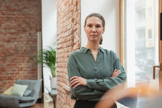 Confident female entrepreneur with arms crossed standing by window in office