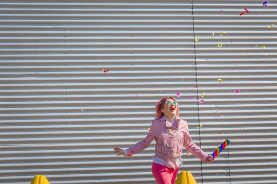Cheerful woman holding party popper while falling confetti in front of metal wall