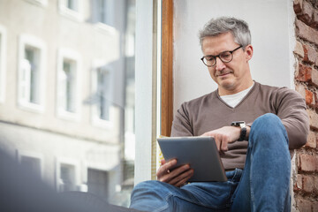 Mature man using digital tablet while sitting on window sill at home