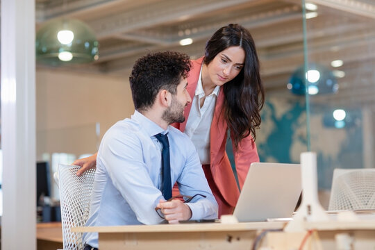 Female Professional Looking At Laptop By Male Colleague At Desk In Coworking Office