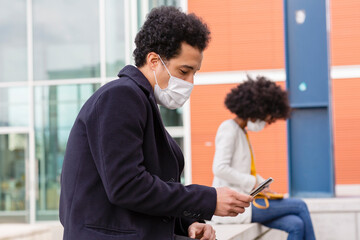 Male professional with protective face mask using smart phone while sitting on retaining wall