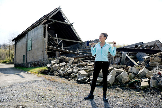 Thoughtful woman standing with hammer on road with destructed house in background