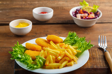 French fries with chicken nuggets next to cheese sauce and ketchup in a gravy boat and salad on wooden planks next to a fork.