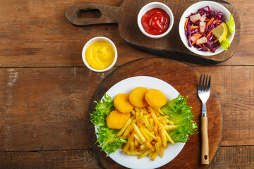 French fries with chicken nuggets next to cheese sauce and ketchup in a gravy boat and salad on wooden planks next to a fork.