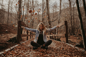 Happy woman playing with autumn leaves while sitting on wooden bridge in forest