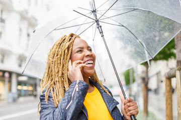 Cheerful woman with braided hair talking on phone while holding umbrella