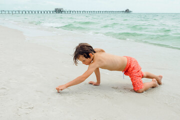 Fototapeta premium Child play on a tropical beach. Kid playing in the sea on summer family vacation. Children have fun at the beach resort. Funny boy writes on the sand.Active children's lifestyle, swimming people