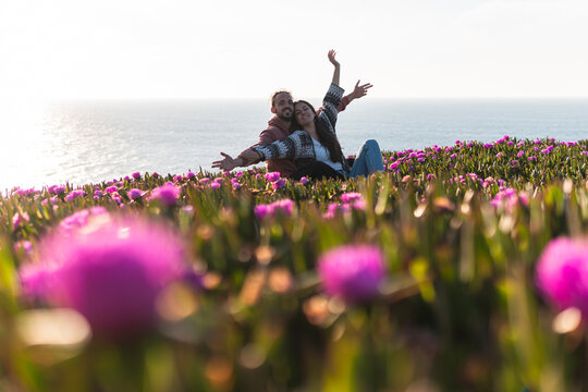 Young couple with arms outstretched sitting at flower filed by sea