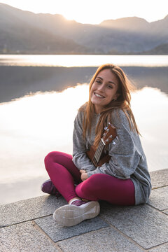 Smiling woman with ukulele sitting cross-legged on promenade near lake