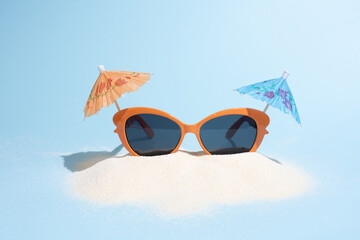 Orange sun glasses with cocktail umbrellas on sand on blue background in sunlight, summer tourism...
