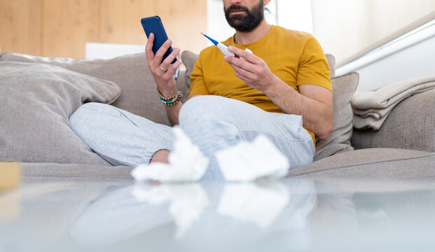 Man holding thermometer and smart phone while sitting on sofa at home