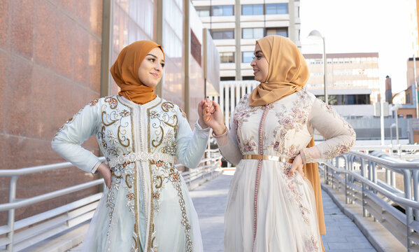 Friends wearing hijab and dress smiling while holding hands on bridge