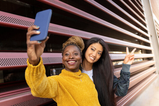 Young woman taking selfie with female friend showing peace sign by wall