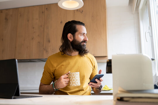 Man with long hair looking away holding smart phone and mug at home