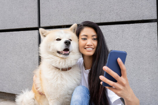 Hipster woman taking selfie with Akita dog through mobile phone by wall