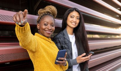 Multi-ethnic female friends looking away holding smart phone outdoors