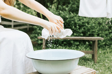 blonde teen girl in linen apron is doing homework washing clothes in basin and hanging them on ropes on the street in courtyard of village cottage house, concept of summer and freshness