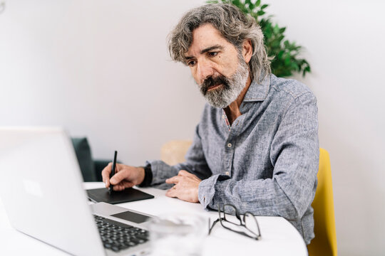 Senior male business professional looking at laptop while using graphics tablet at home office