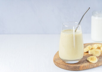 Indian lassi drink or milkshake made of ripe sweet bananas blended with milk served in drinking glass with metal straw and fruit slices on cutting board on white wooden table. Image with copy space