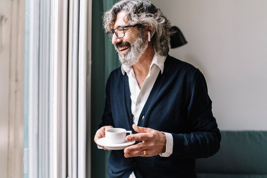 Happy senior man with in-ear headphones holding coffee cup near window at home