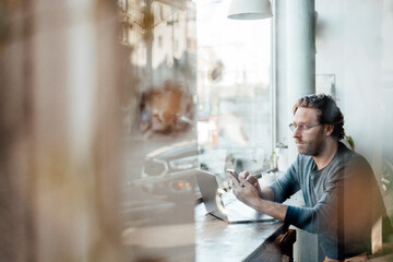 Mature man text messaging on mobile phone by laptop at coffee shop