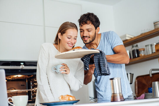 Man showing freshly baked croissant to girlfriend in kitchen at home