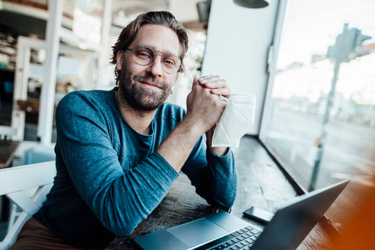 Smiling confident man by laptop in coffee shop during COVID-19