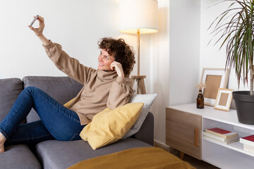 Cheerful woman taking selfie on smart phone while sitting on sofa at home