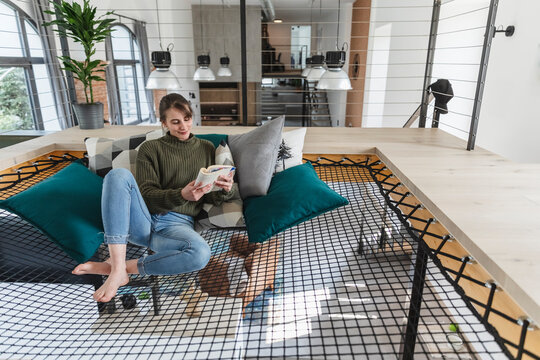 Woman reading book while sitting on hammock in modern home