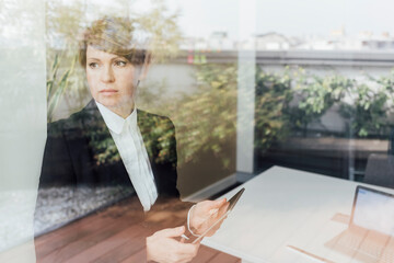 Serious businesswoman holding mobile phone while looking through window in office